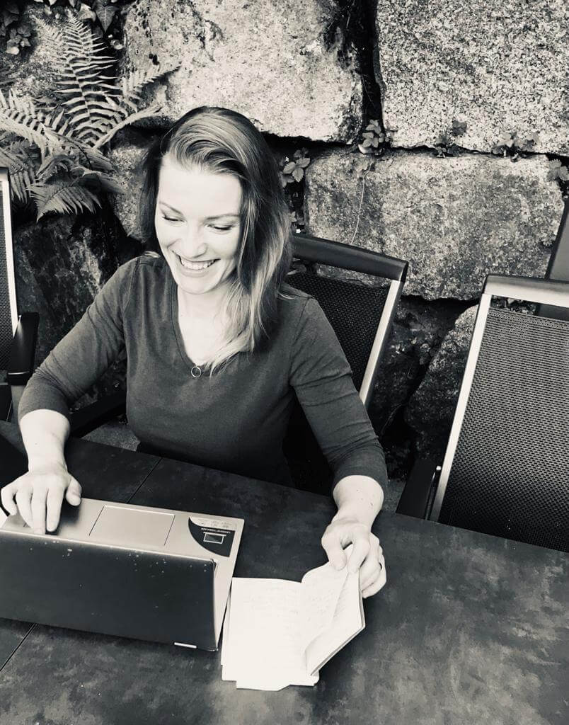 Schwarzweiß-Bild der Autorin Noa Arden bzw. Sarah Maynight tippt am Laptop und lächelt.