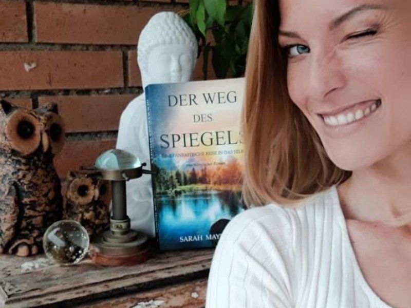 Autorin Sarah Maynight bzw. Noa Arden zwinkert, im Hintergrund eine weiße Buddha Figur, Zwei Toneulen, ein Kompass und das Buch Der Weg des Spiegels